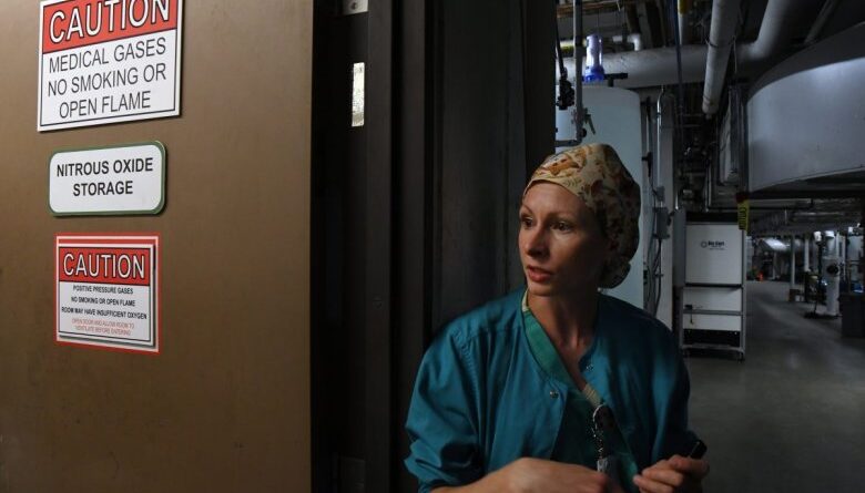 A man in scrubs and a cap stands by a door with signs warning of medical gases, no smoking, and nitrous oxide storage in a dimly lit industrial hallway.